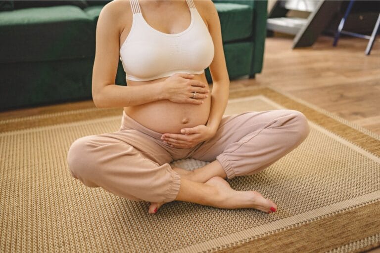 Pregnant woman sitting cross-legged on a woven rug, cradling her belly with both hands, wearing a white sports bra and beige pants.