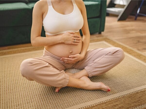 Pregnant woman sitting cross-legged on a woven rug, cradling her belly with both hands, wearing a white sports bra and beige pants.